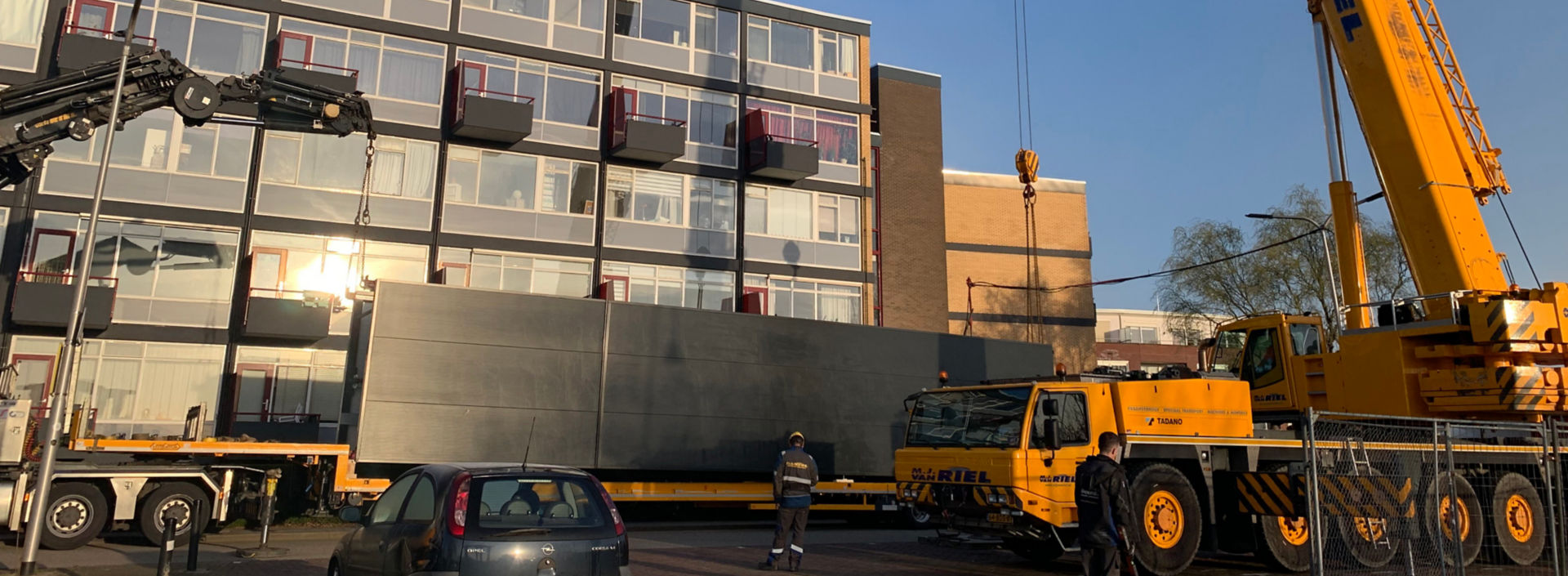 Crane getting ready to lift a prefabricated elevator from a truck in front of a residential building.
